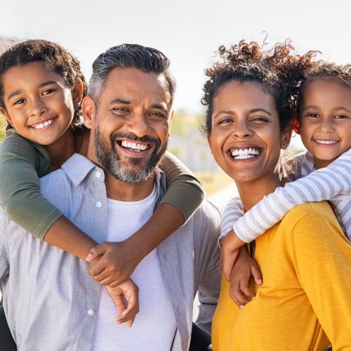 Mixed race parents giving piggyback ride to their children outdoor. Portrait of happy african mother and indian father with daughters looking at camera outside house. Smiling and joyful family standing with their beautiful little girls.